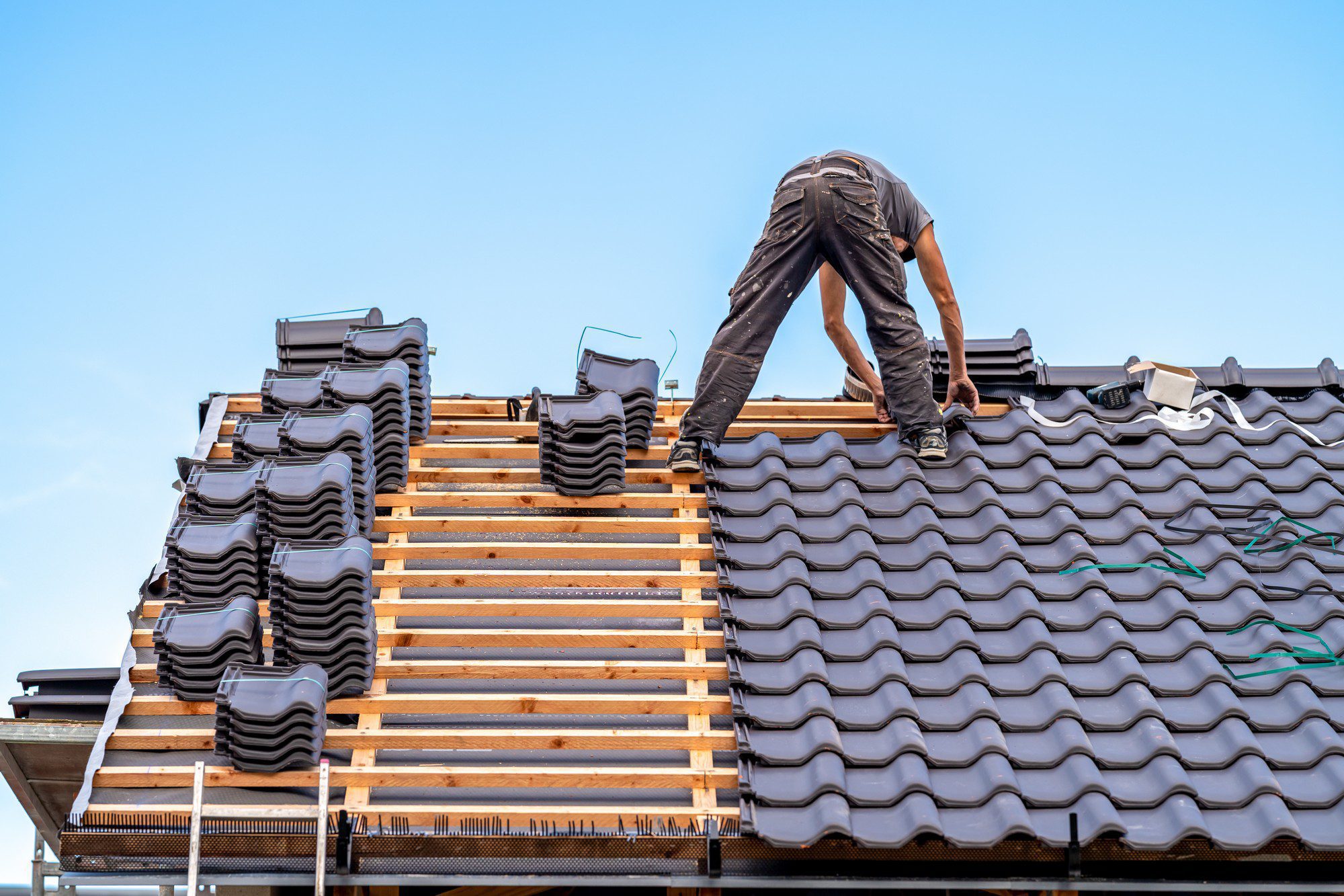 The image shows a person working on a roof. They appear to be in the process of installing roofing tiles. The wooden roof structure is visible, with battens in place to support the tiles. Several stacks of black roof tiles are neatly piled on the roof, and some tools and materials are scattered around the work area. The person is bending over, likely adjusting or fixing a tile in place. The sky is clear, indicating good weather for outdoor construction work. Safety precautions, such as the proper footwear and gloves, seem to be taken by the worker.