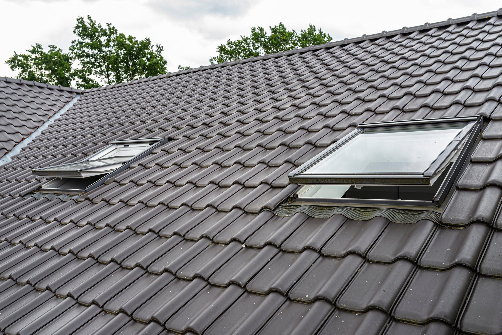 The image shows a close-up of a house roof. The roof is covered with dark grey interlocking tiles, a common roofing material. There are two roof windows, also known as skylights, installed on the slope of the roof, which allow natural sunlight to enter the space beneath the roof, commonly used to light up attic or loft spaces. The roof also has a ridgeline at the top, with trees and a cloudy sky visible in the background.